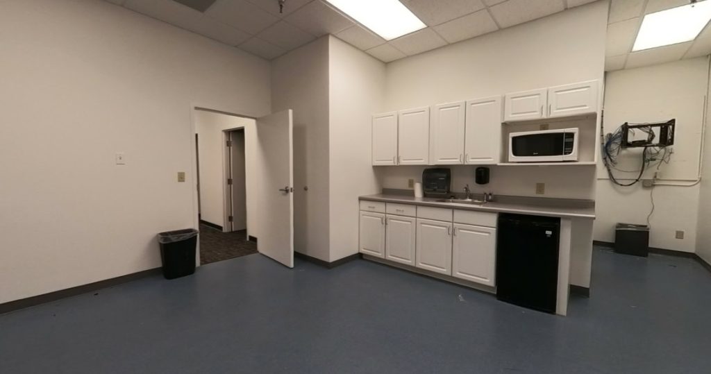 Small kitchenette with white cabinets, sink, microwave, mini fridge, and a trash can. The walls are white, the floor is blue, and one door is open to an adjacent room in Suite 100 at Kubias.