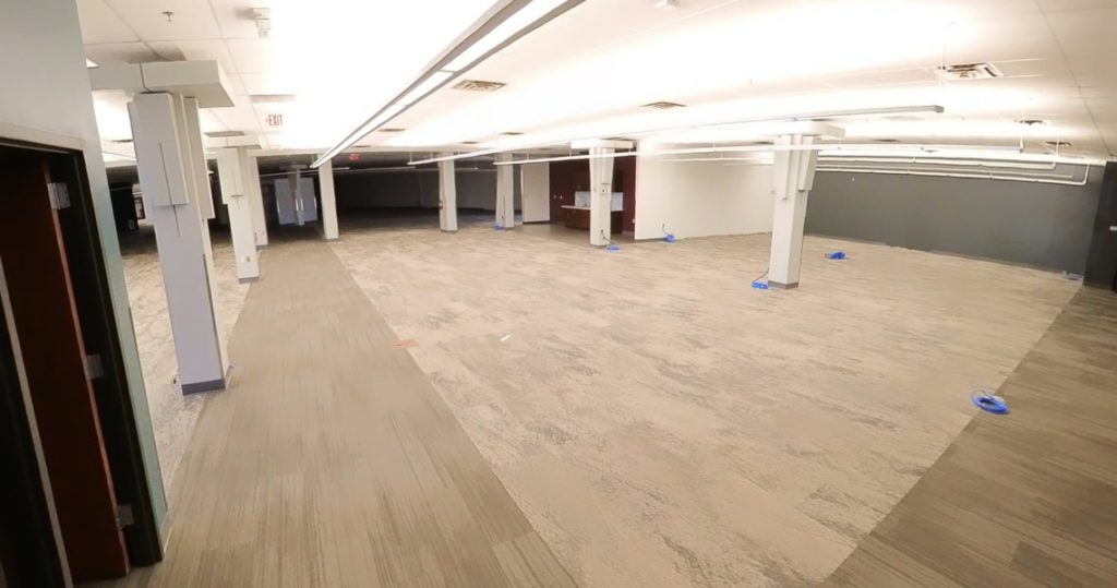 A wide, empty office space at 3281 Armar Drive features columns, overhead lights, and a mix of beige and gray carpeting; blue tape marks are on the floor.