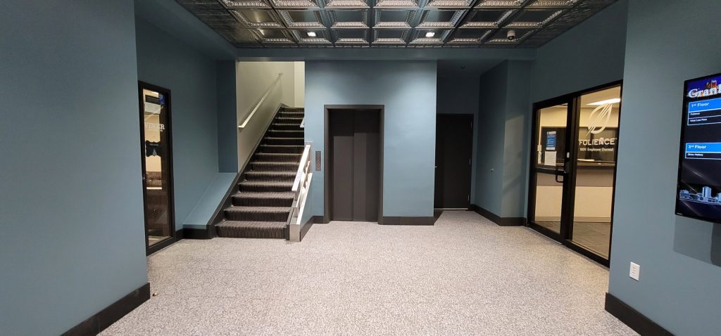 A hallway in the Granby Building with gray walls features a staircase on the left, an elevator in the center, and office doors with glass windows on both sides.
