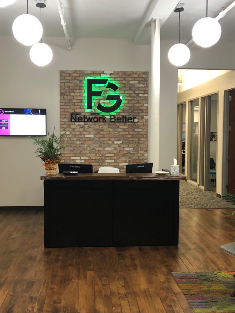 Reception area with a wooden desk, two chairs, and a brick wall featuring the green FG Network Better logo. Light fixtures hang from the ceiling and a monitor on the left wall highlights our latest Tenant Spotlight.