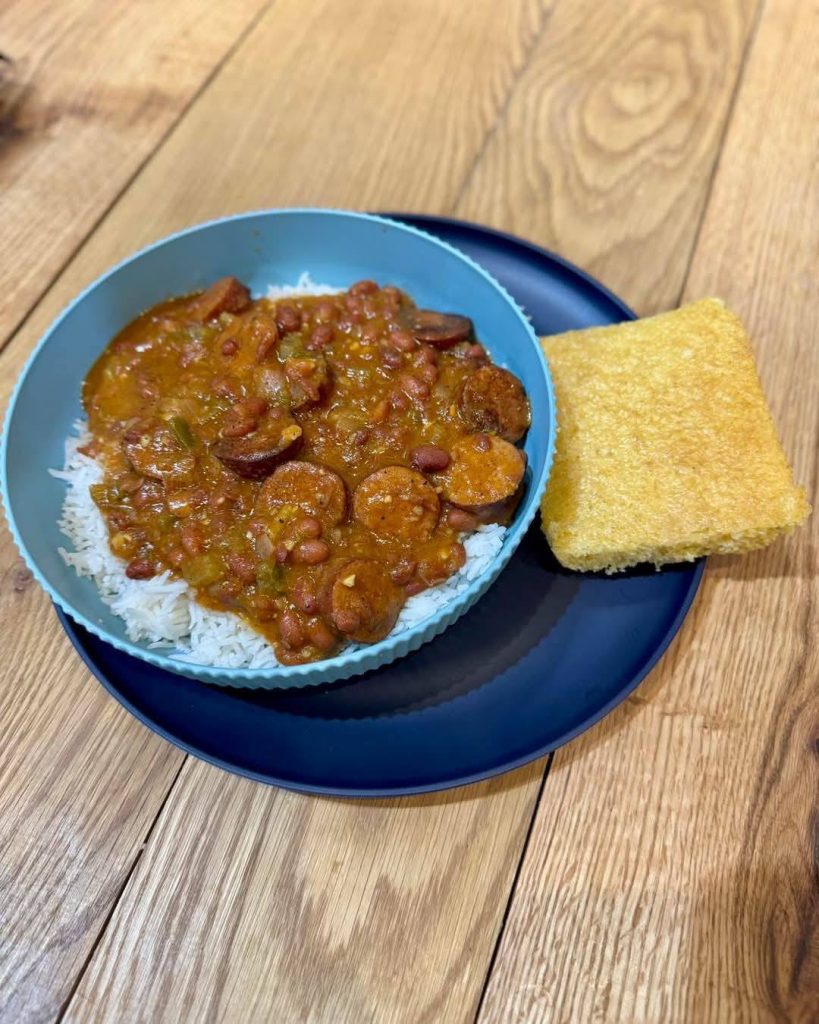A bowl of Cajun rice topped with sausage and bean stew, served with cornbread on a blue plate at KC Cakes in Downtown Cedar Rapids, placed on a wooden table.