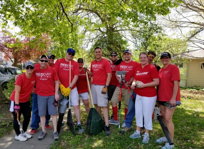 A group of people wearing matching red REDDAY shirts pose outdoors with gardening tools under a tree, participating in a Keller Williams volunteer event.