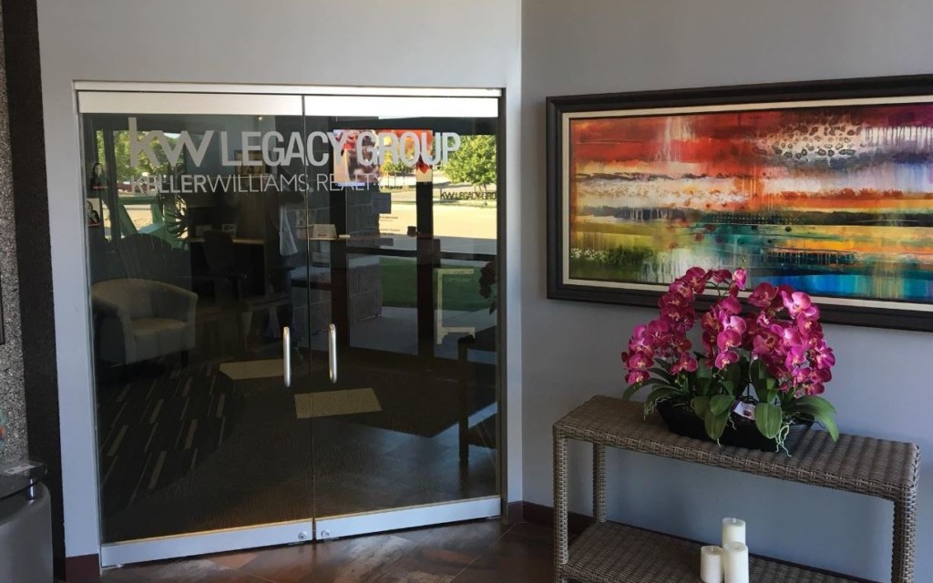 Glass doors labeled KW LEGACY GROUP KELLER WILLIAMS REALTY, a leading realty company, stand beside a table with purple orchids, candles, and a colorful abstract painting on the wall.