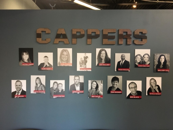 A wall display labeled Cappers features a Tenant Spotlight with headshots of individuals arranged in two rows, each labeled with names and some photos featuring red accents, highlighting achievements within the Keller Williams realty company.