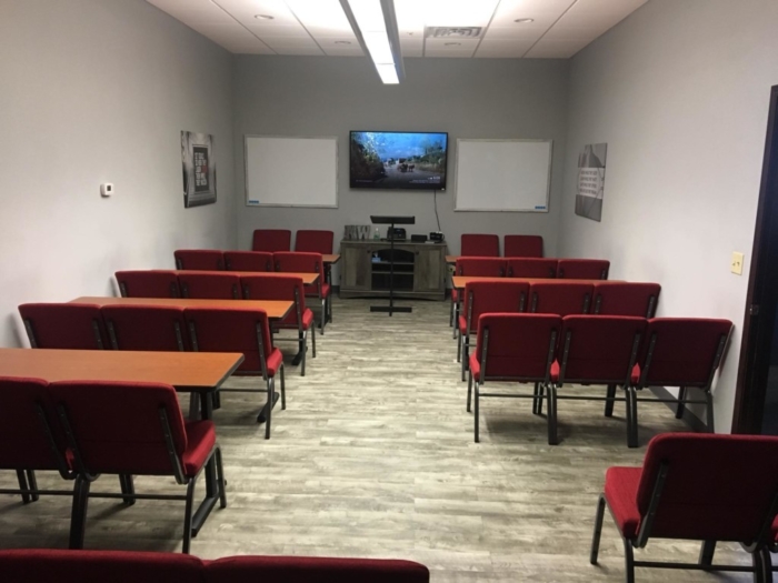Classroom with red chairs and wooden tables facing a TV screen, two whiteboards, and a lectern at the front; walls are gray with framed posters featuring Keller Williams Realty, our featured Tenant Spotlight realty company.