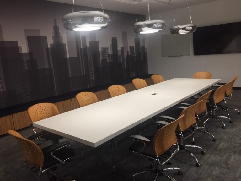 A modern conference room with a long white table, ten brown chairs, overhead lights, a cityscape mural, a wall-mounted TV, and a whiteboard.