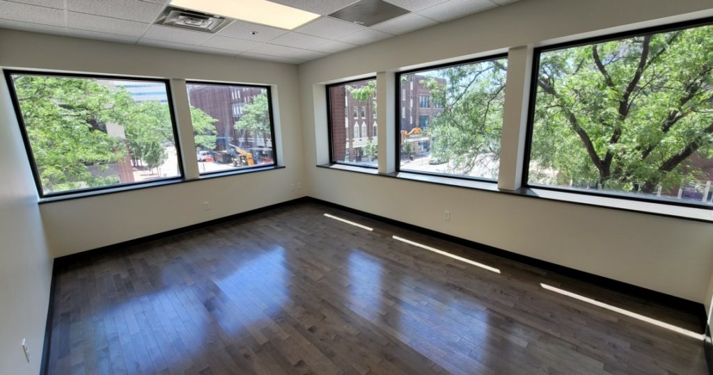 Sunlight pours into Suite 200 of the Granby Building, an empty office room featuring dark wood floors, crisp white walls, and large windows that reveal vibrant trees and city buildings outside.