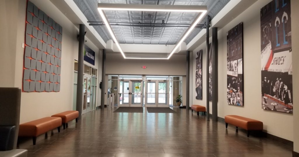 A modern building lobby at the Armstrong Centre features brown benches, wall art, and a square ceiling light fixture, leading to glass double doors at the far end.