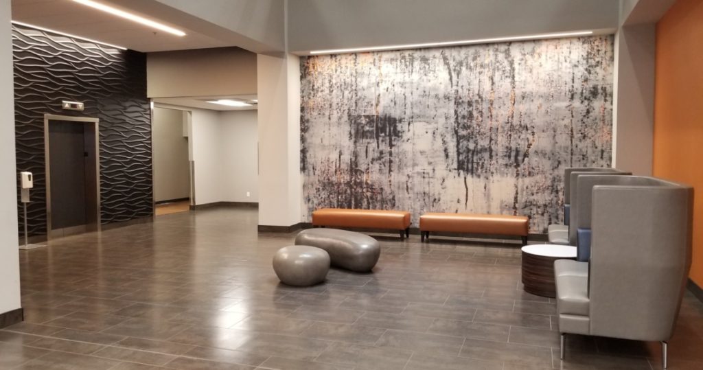 Modern lobby at Armstrong Centre with abstract wall art, orange bench seating, two gray sculptural stools, high-backed chairs, tiled floor, and an elevator in the background.