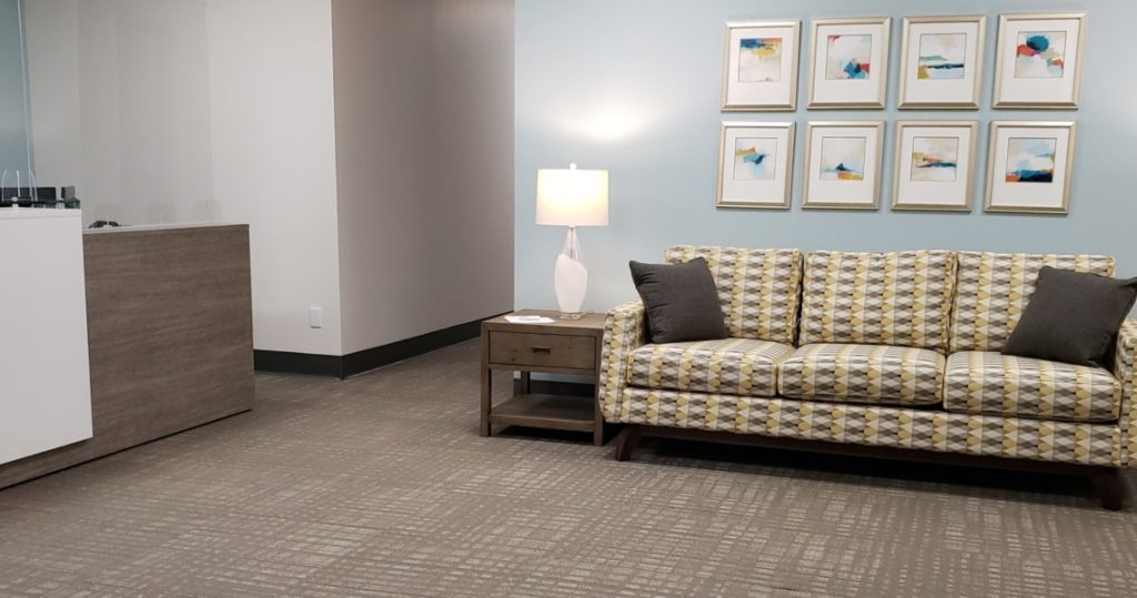 A modern waiting area at Armstrong Centre features a patterned sofa, two dark cushions, a side table with a lamp, and six abstract pictures on a light blue wall.