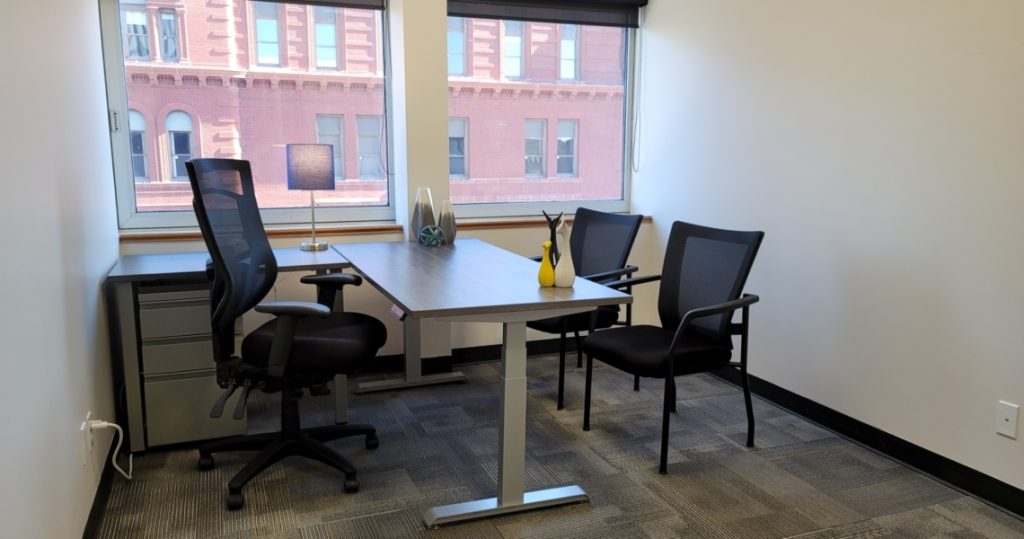 Small office space with a desk, three black chairs, a lamp, decorative vases, and two windows showing a brick building outside—ideal for those seeking professional office suites at Armstrong Office Suites.