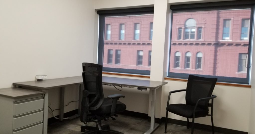 Two black office chairs and a gray desk in a small Armstrong Office Suites space with two large windows overlooking a red brick building.