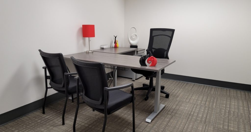 Modern office space with a desk, mesh chairs, a red lamp, headphones, and abstract decor on a beige carpeted floor against a plain white wall—perfect for the professional atmosphere found in Armstrong Office Suites.