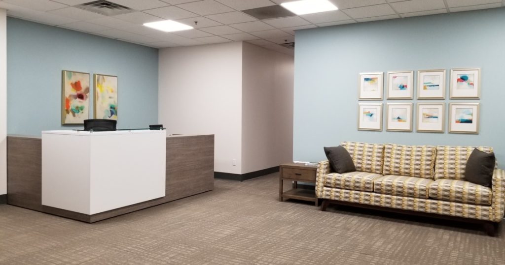 Reception area with a modern desk, patterned sofa, side table, and abstract artwork on pale blue and white walls under a grid ceiling—an inviting entrance that reflects the contemporary style of Armstrong Office Suites.