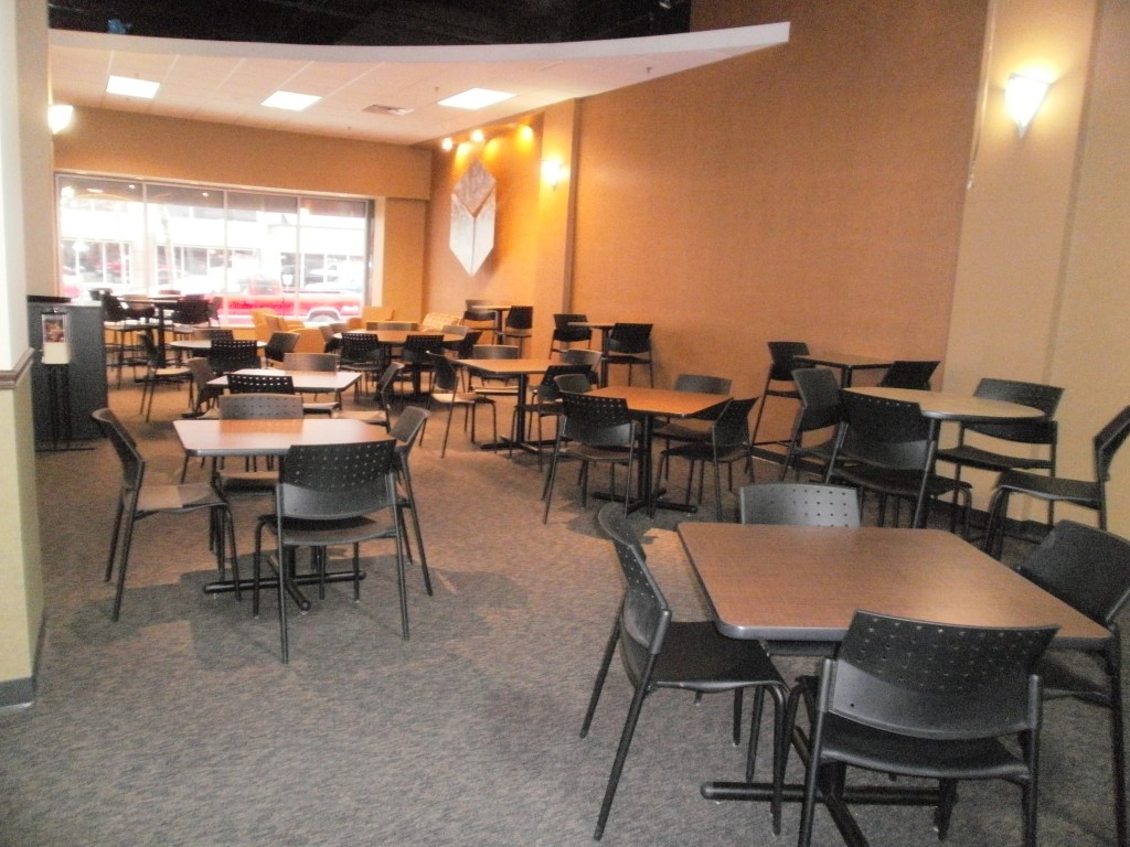 A cafeteria in the Barnes-Chamberlain Building with empty tables and chairs, beige walls, a carpeted floor, and large front windows letting in natural light.