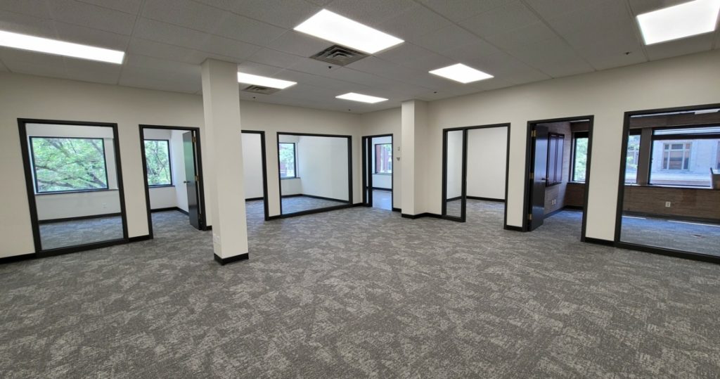 Empty office space with gray carpet, white walls, and glass-walled private offices along the perimeter in the Granby Building. Ceiling lights are on, and windows in Suite 200 offer views of trees and buildings outside.