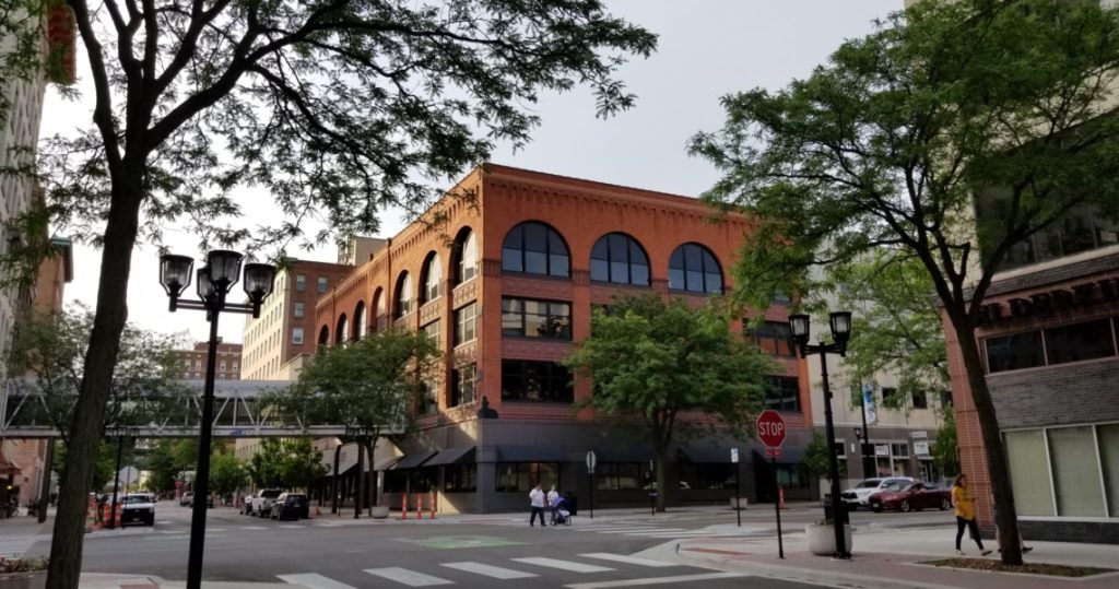 The Granby Building, a red-brick structure with arched windows, stands at a tree-lined street corner in an urban area, with people walking on the sidewalk and cars parked nearby. Suite 400 is located within this vibrant setting.