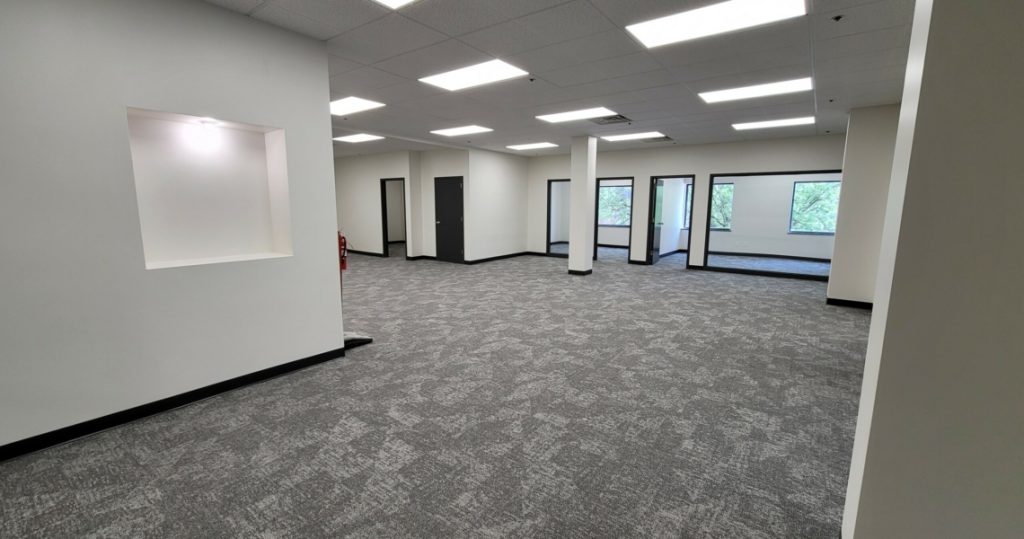 Spacious, empty office in Suite 200 of the Granby Building featuring gray carpet, white walls, ceiling lights, several doors, and windows overlooking greenery.
