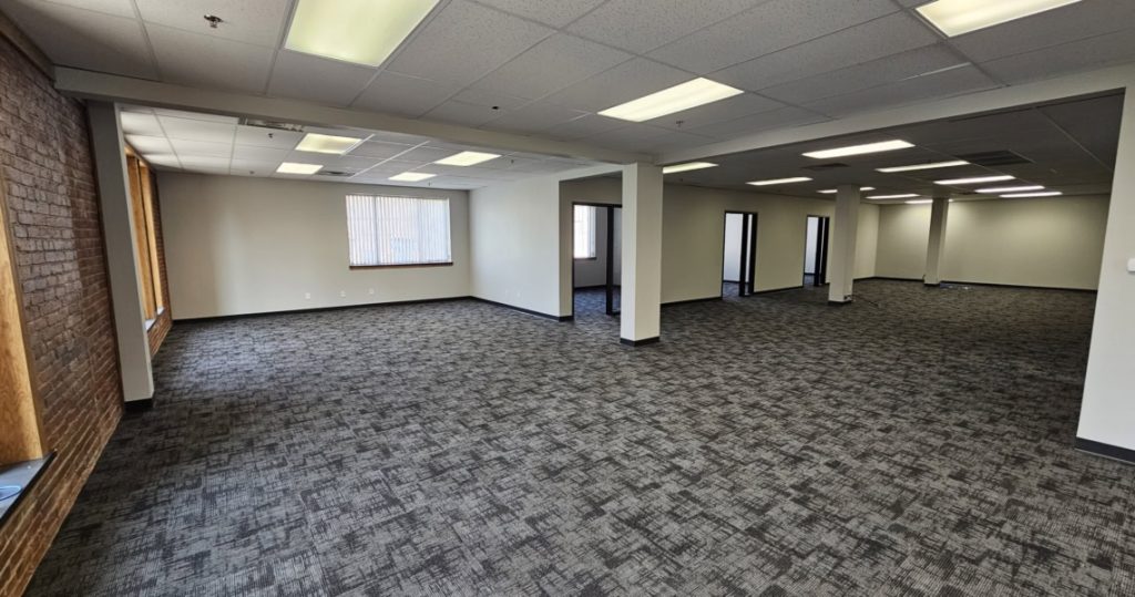 Large, empty office space with gray carpet, white walls, fluorescent lighting, and several windows along one side. Multiple doorways lead to separate rooms in the back, all located in Suite 300 at Kubias.
