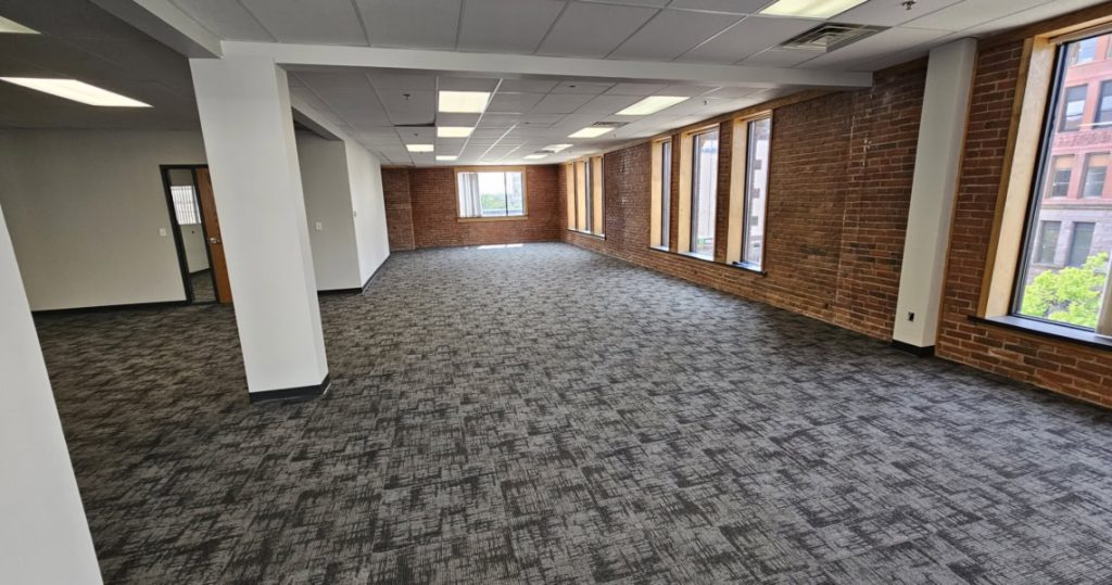 Suite 300 offers an empty office space featuring gray carpet, white walls, exposed brick walls, large windows, and overhead fluorescent lighting—an ideal blank canvas for your business at Kubias.