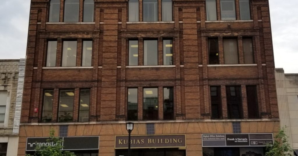 A four-story brick building with large windows, labeled Kubias Building above ground-level shops, is shown on an overcast day. The third floor houses Suite 305, adding to the commercial charm of the historic Kubias landmark.