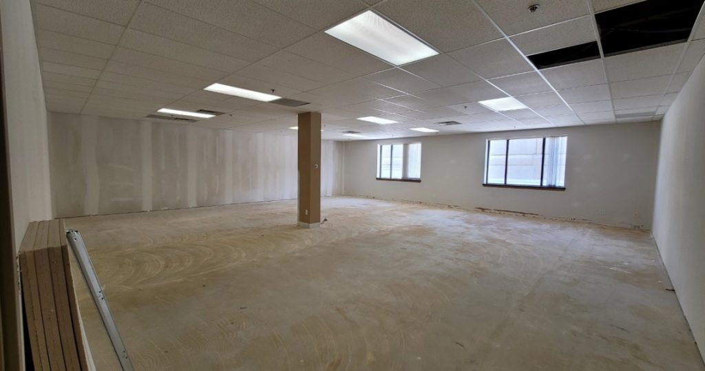 Suite 305 features an empty room with unfinished walls, exposed flooring, and ceiling tiles removed near the windows, all illuminated by fluorescent lights and natural light streaming through two windows—a distinctive space within the Kubias building.