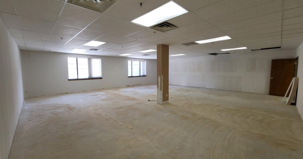 Spacious and versatile, Suite 305 offers an empty office space with bare floors, white walls, a central column, drop ceiling, fluorescent lights, two windows, and a closed door—an ideal blank canvas for your business at Kubias.