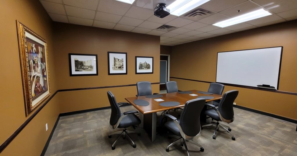 A small BC Office Suites conference room with six chairs around a wooden table, framed pictures on brown walls, and a whiteboard at the front.