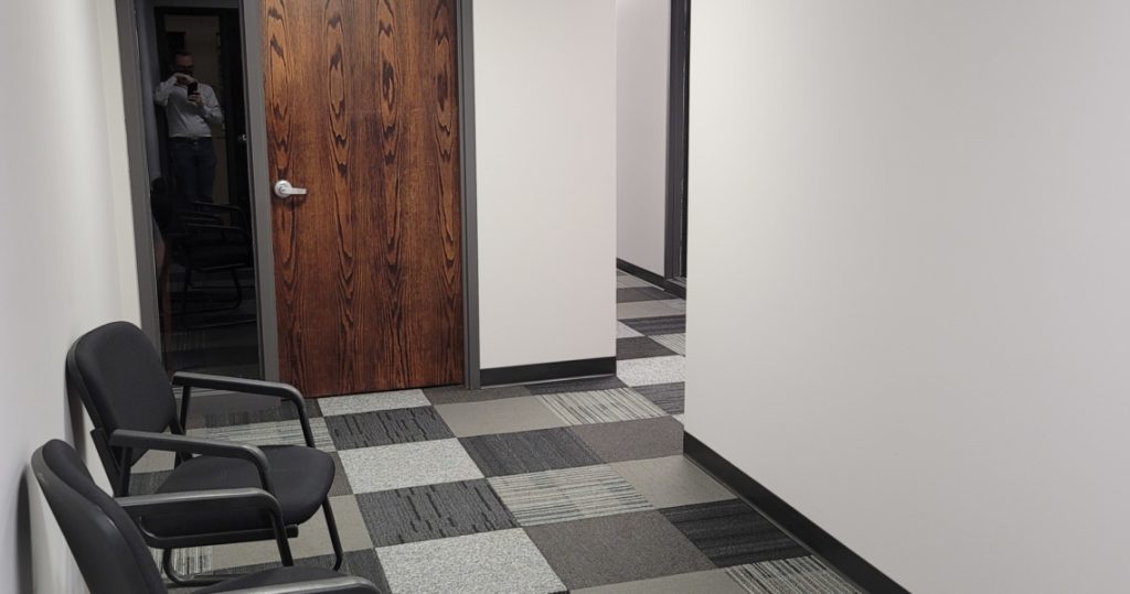 A hallway in BC Office Suites with two black chairs against a white wall, a closed wooden door, and patterned carpet tiles; a person is reflected in the glass next to the door.