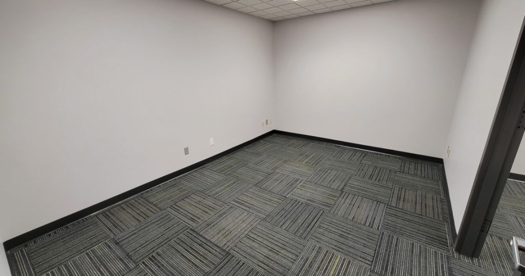 Empty office room with white walls, gray patterned carpet, and visible electrical outlets along the baseboards. The ceiling features a grid pattern with recessed lighting—ideal for your business at BC Office Suites.