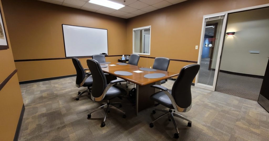 Conference room at BC Office Suites with six office chairs around a square table, placemats on the table, a whiteboard on the wall, and a window to the hallway.