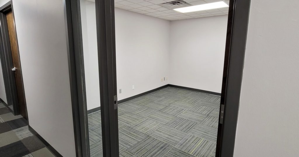 A small, empty BC Office Suites room with grey patterned carpet, white walls, and a drop ceiling, viewed through an open doorway from a hallway.