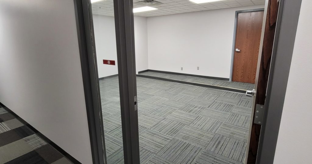 A small, empty BC Office Suites room featuring gray patterned carpet, white walls, a wooden door, and a glass partition.
