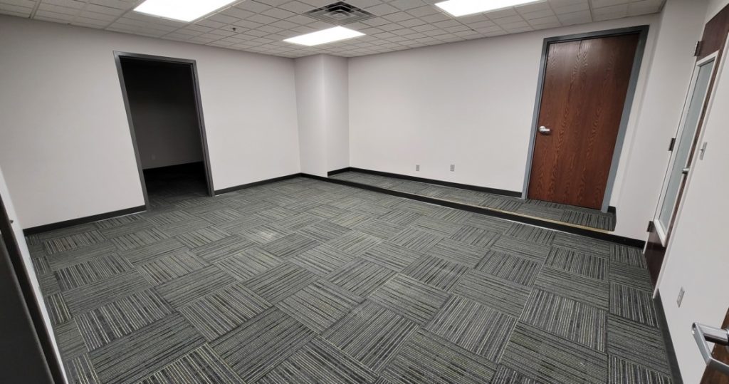 Empty BC Office Suites room with gray patterned carpet, white walls, a wooden door, an open doorway, and a lowered section of floor near the back wall.